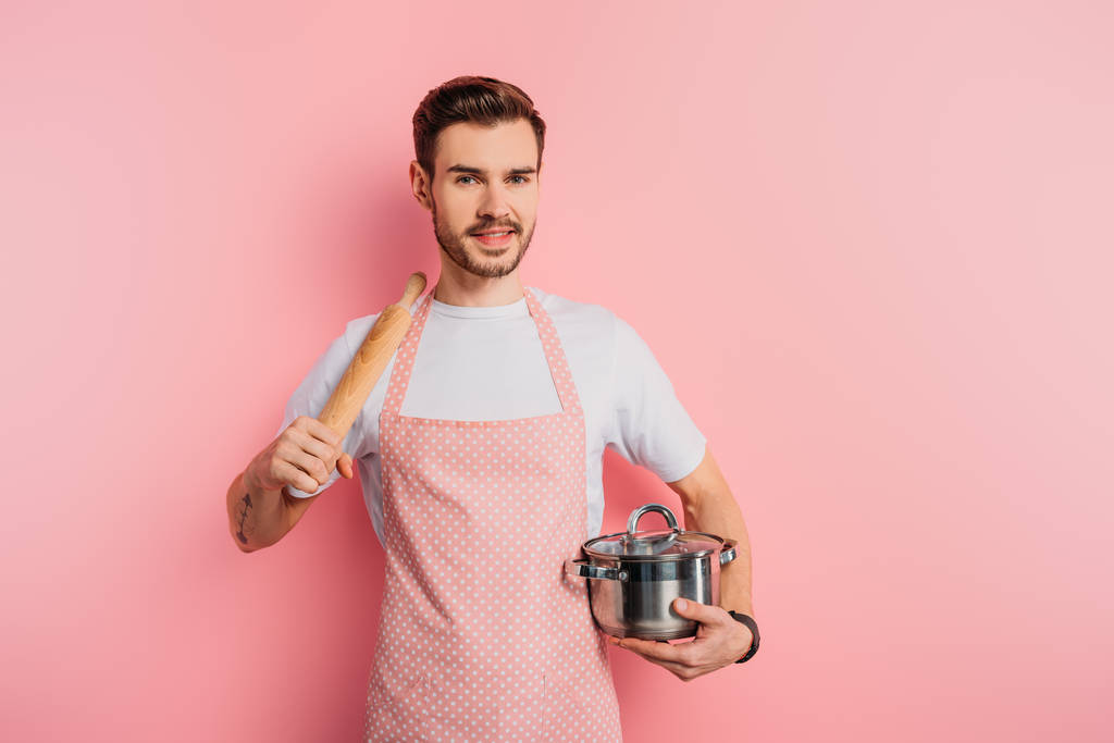 cheerful young man in apron holding saucepan and rolling pin on pink background  - Photo, Image