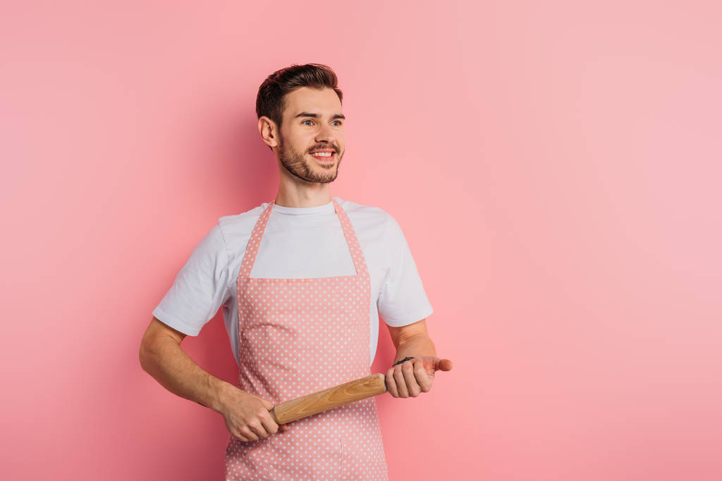 cheerful young man in apron looking away while holding rolling pin on pink background - Photo, Image