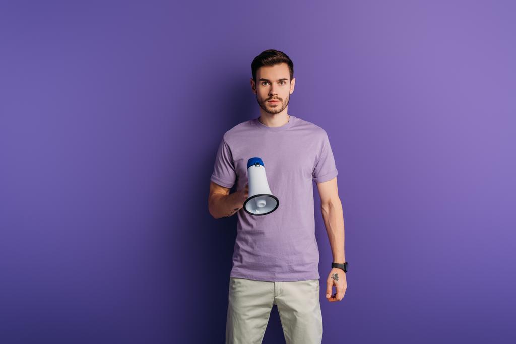 seriózní mladý muž drží megafon a dívá se na kameru na fialovém pozadí - Fotografie, Obrázek