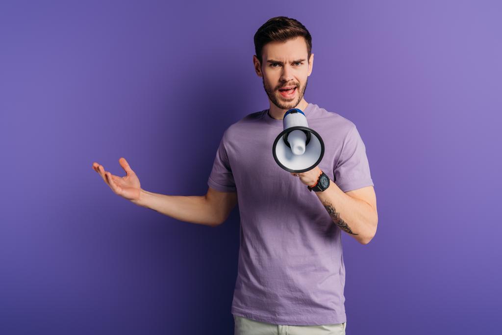 displeased young man screaming in megaphone while standing with open arm on purple background - Photo, Image