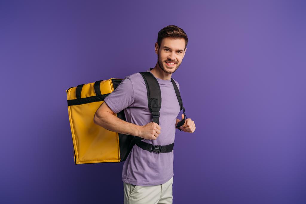 szczęśliwy dostawca patrząc w kamerę podczas noszenia termo plecak izolowany na fioletowy - Zdjęcie, obraz