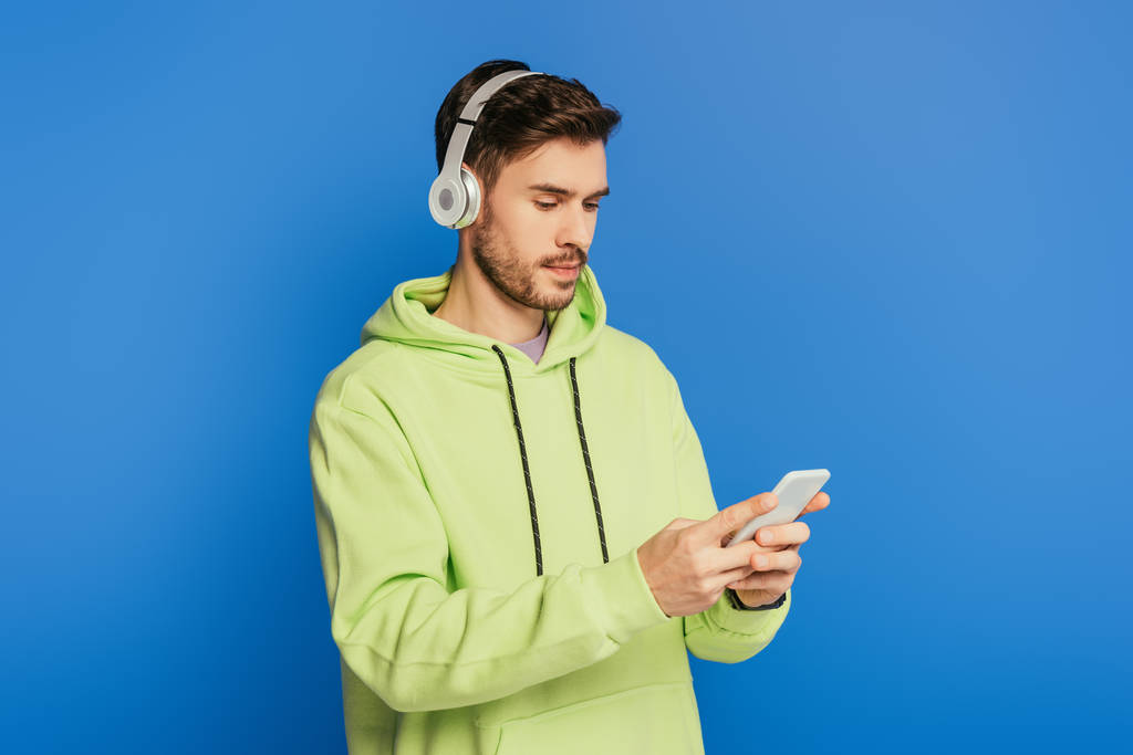 Kablosuz kulaklıklı dikkatli genç adam mavi üzerinde izole akıllı telefon kullanıyor. - Fotoğraf, Görsel