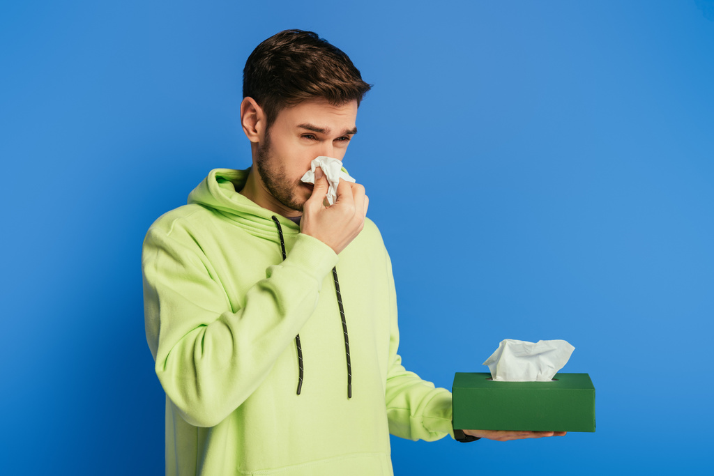 joven enfermo limpiando la nariz con servilleta de papel sobre fondo azul
 - Foto, Imagen