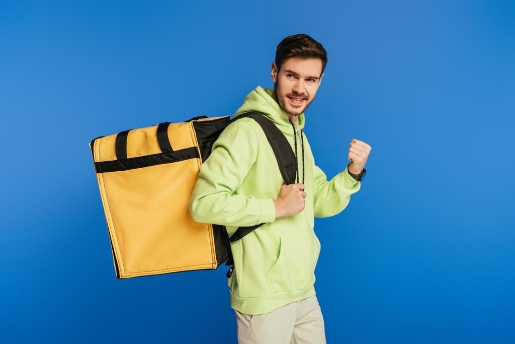 happy delivery man showing winner gesture while carrying thermo backpack isolated on blue - Photo, Image