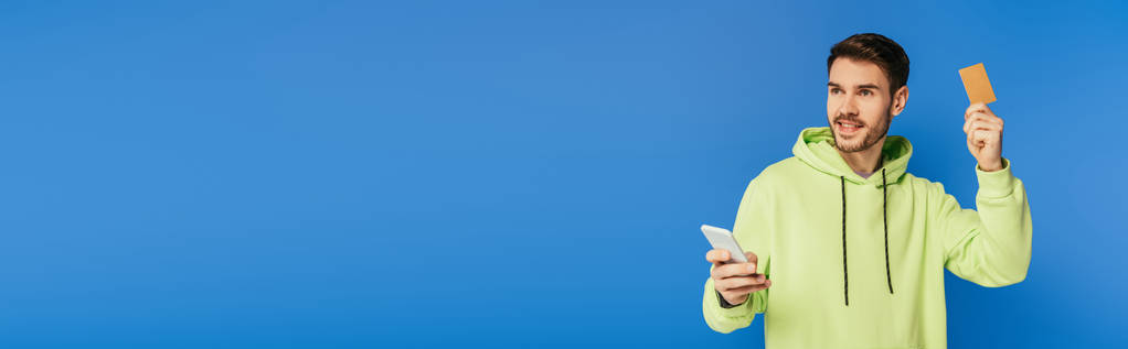 panoráma lövés mosolygós férfi gazdaság okostelefon és hitelkártya elszigetelt kék - Fotó, kép