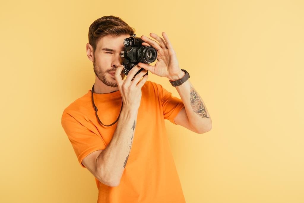 attentive handsome photographer taking picture with digital camera isolated on yellow - Photo, Image