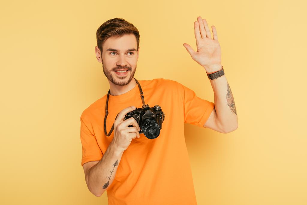 Sarı arka planda el sallayan dijital kameralı yakışıklı, neşeli fotoğrafçı. - Fotoğraf, Görsel