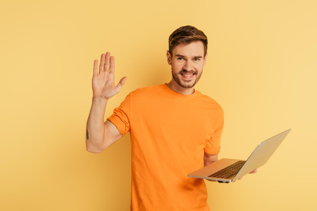 ευτυχισμένος άνθρωπος κουνώντας το χέρι στην κάμερα, ενώ κρατώντας φορητό υπολογιστή σε κίτρινο φόντο - Φωτογραφία, εικόνα