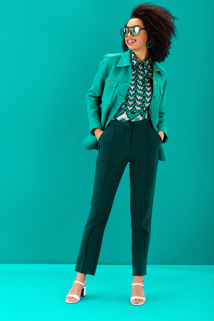 sonriente mujer afroamericana en chaqueta elegante con las manos en los bolsillos sobre fondo turquesa
  - Foto, imagen