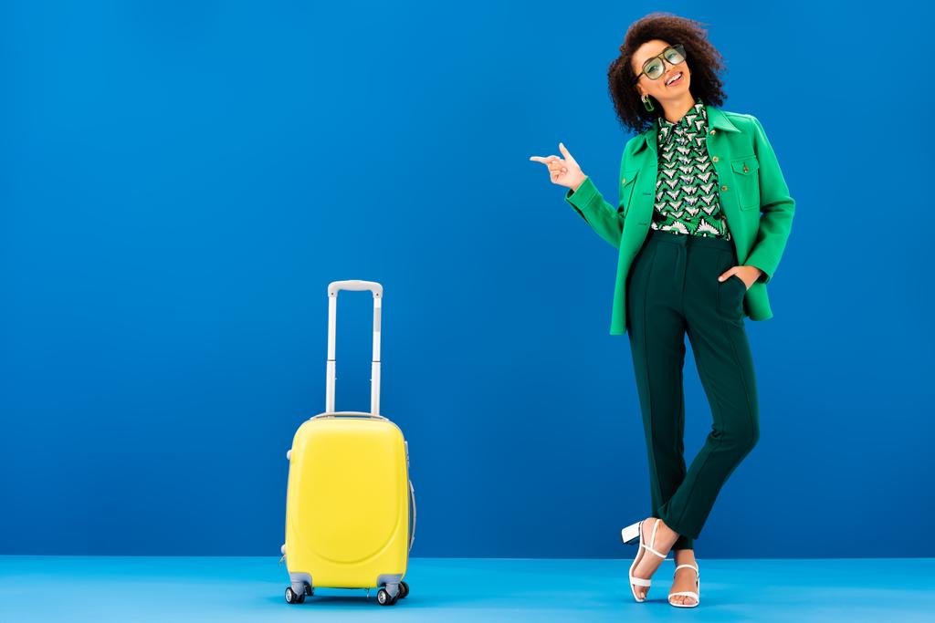 アフリカ系アメリカ人の女性が指を指して青い背景に旅行バッグの近くに立ち - 写真・画像