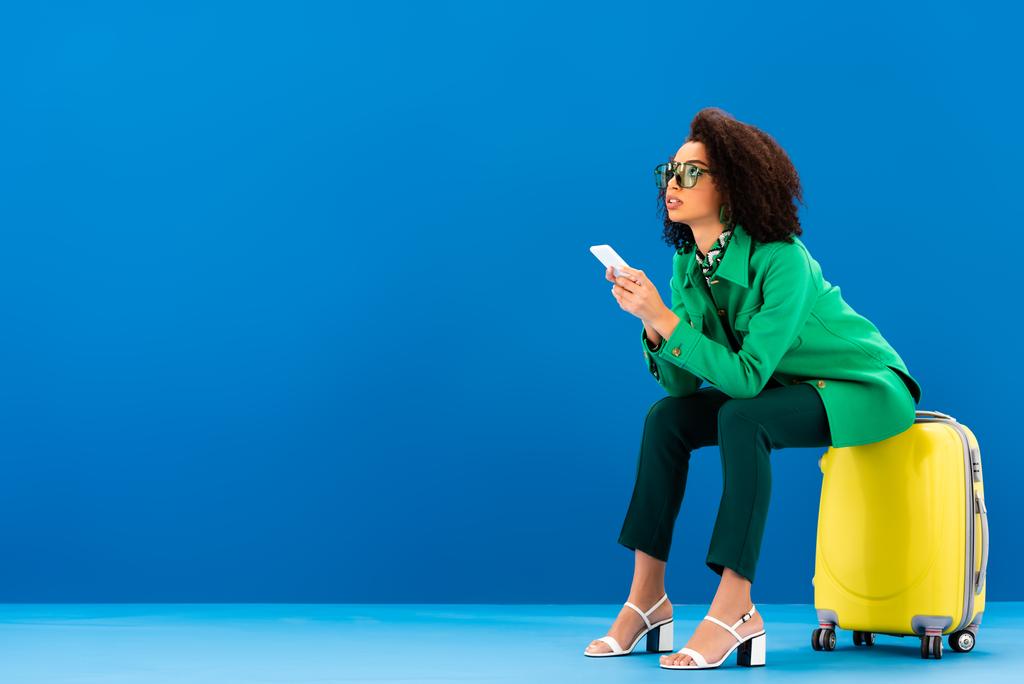 african american woman sitting on travel bag and holding smartphone on blue background  - Photo, Image