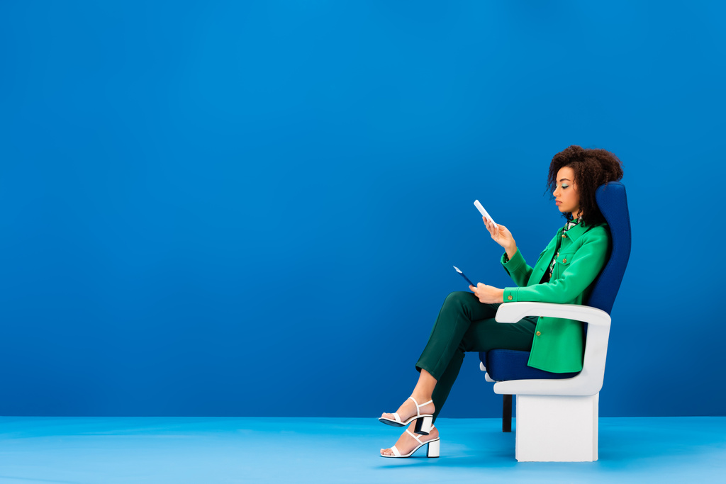 vista lateral da mulher americana africana sentado no assento e segurando smartphone no fundo azul
  - Foto, Imagem