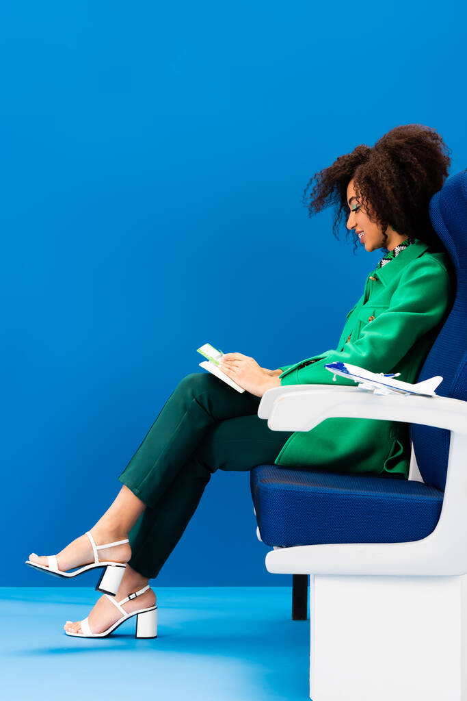 πλαϊνή άποψη του χαμογελαστού Αφροαμερικανός γυναίκα και το μοντέλο του αεροπλάνου στο κάθισμα σε μπλε φόντο  - Φωτογραφία, εικόνα