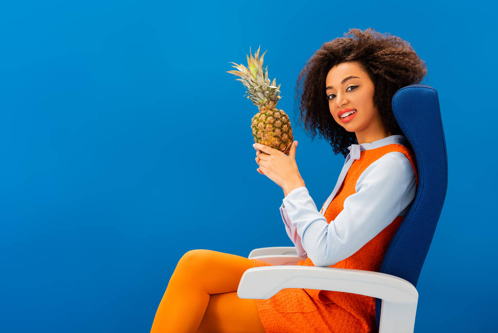 usmívající se africký Američan v retro šaty sedí na sedadle a drží ananas izolované na modré  - Fotografie, Obrázek