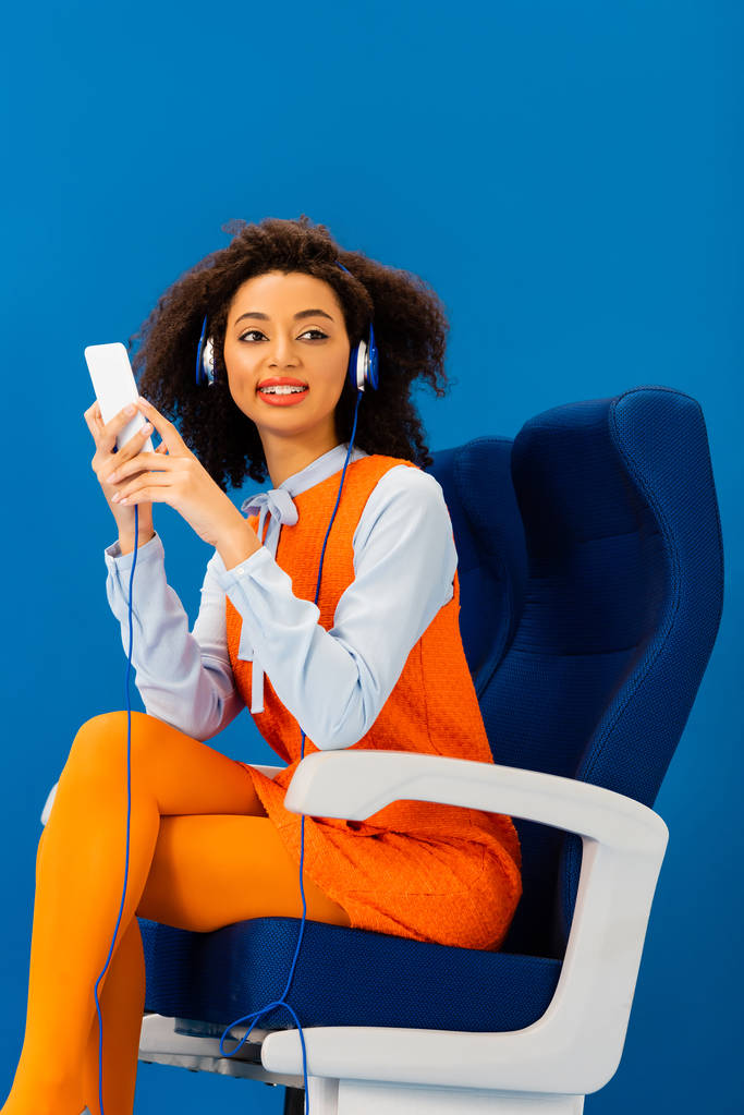 усміхнений афроамериканець в ретро-сукні слухає музику і використовує смартфон ізольовано на синьому
  - Фото, зображення