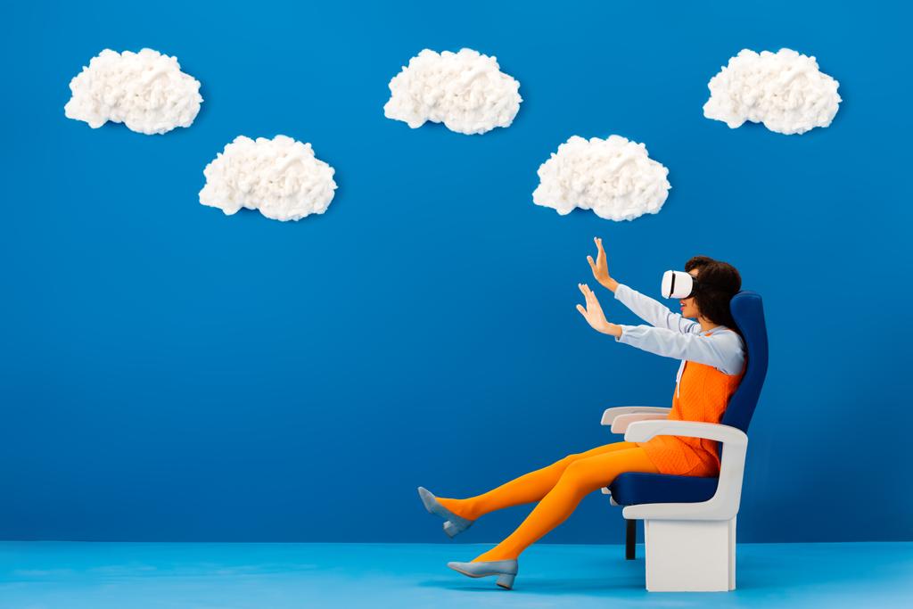 vista lateral de afroamericano en auriculares vr con las manos extendidas sentadas en el asiento sobre fondo azul con nubes
  - Foto, imagen