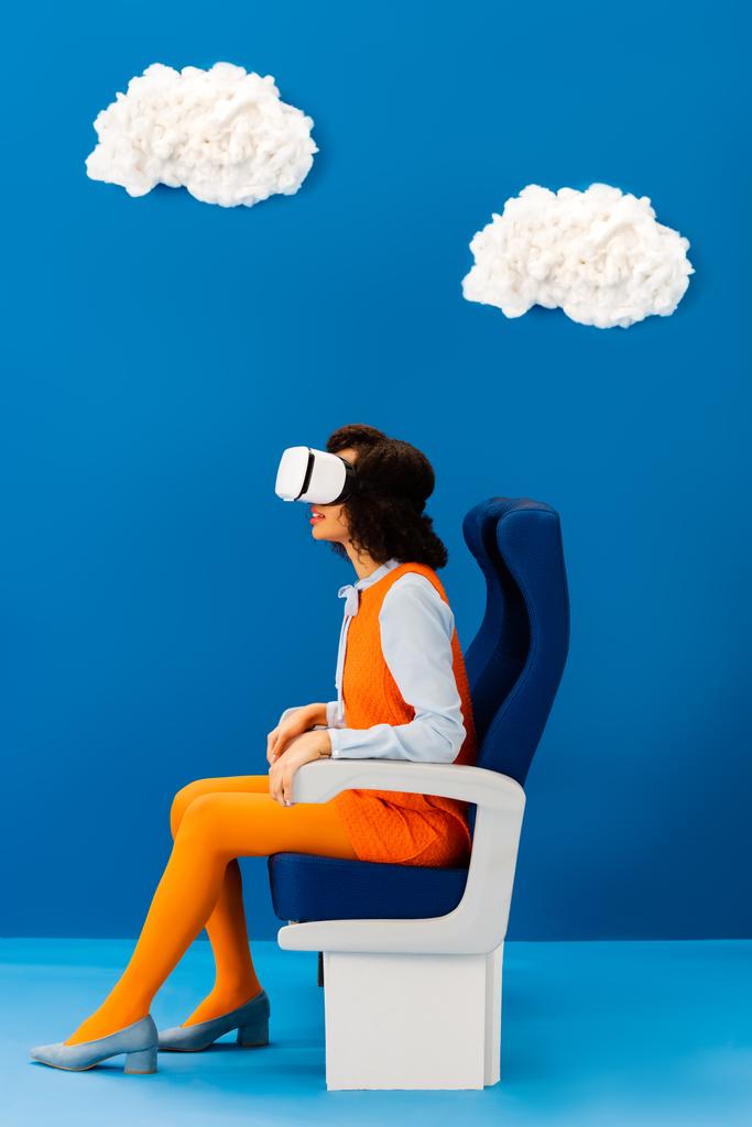 Seitenansicht des Afroamerikaners in vr Headset sitzt auf Sitz auf blauem Hintergrund mit Wolken  - Foto, Bild