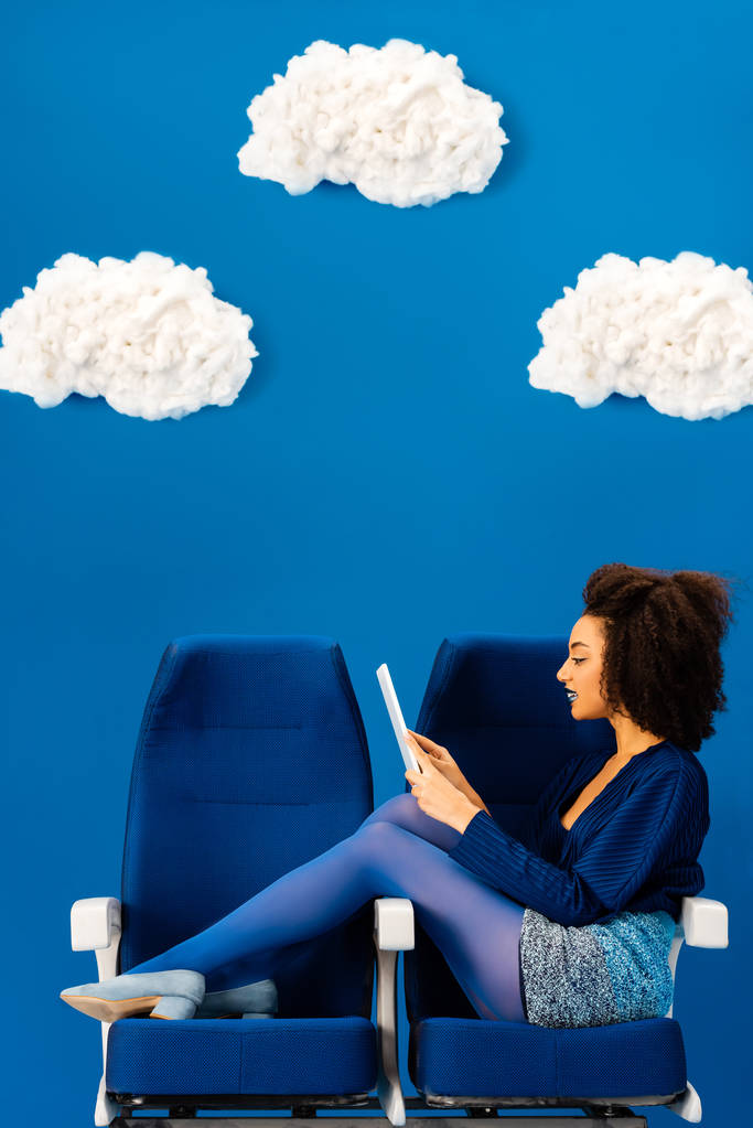 vista laterale di afro americano seduto sui sedili e utilizzando tablet digitale su sfondo blu con nuvole
  - Foto, immagini