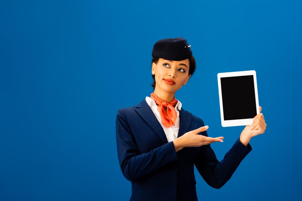 smiling african american flight attendant pointing with hand at digital tablet isolated on blue  - Photo, Image