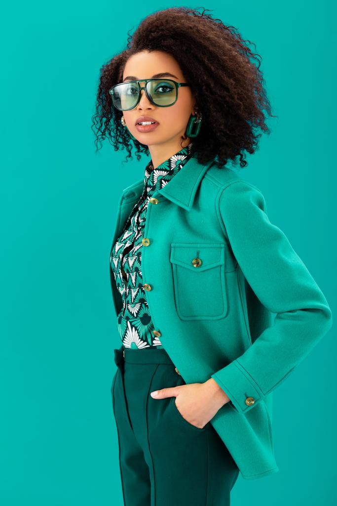 stylish african american woman with hand in pocket looking at camera isolated on turquoise - Photo, Image