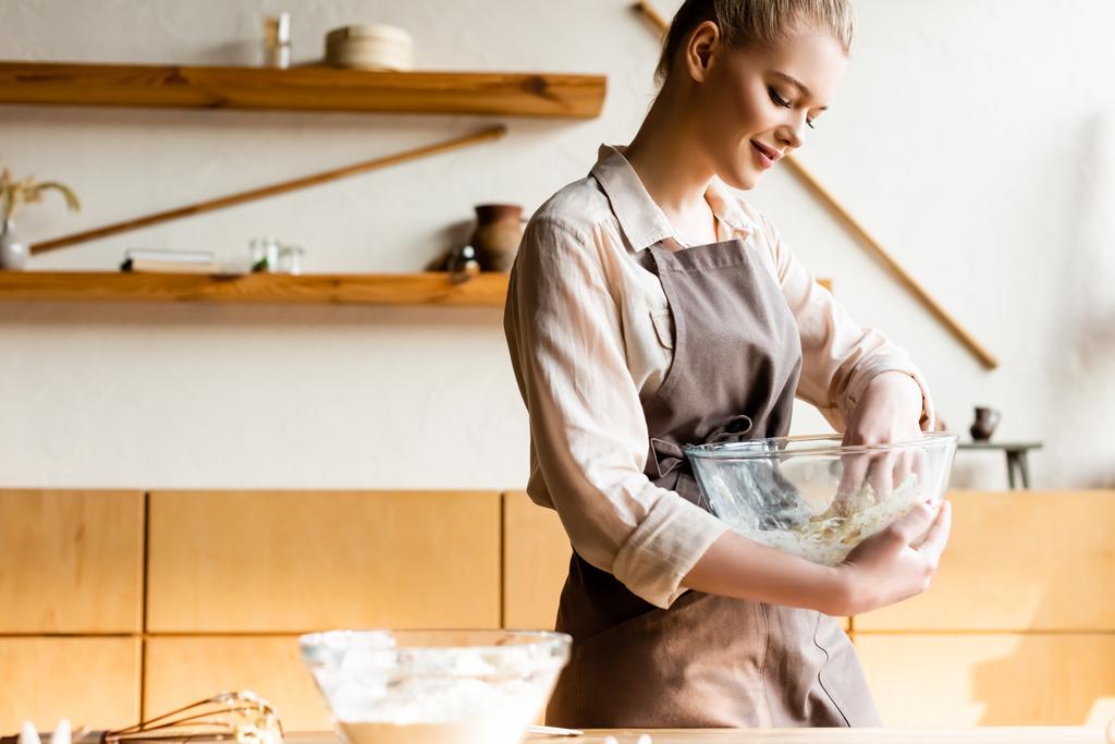 happy woman in apron holding bowl while kneading dough  - Photo, Image