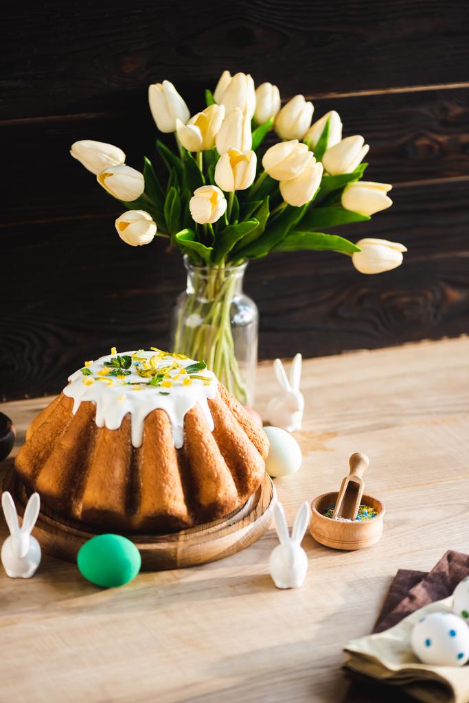 foco seletivo de delicioso bolo de Páscoa perto de estatuetas com coelhos de Páscoa, ovos pintados e tulipas
  - Foto, Imagem