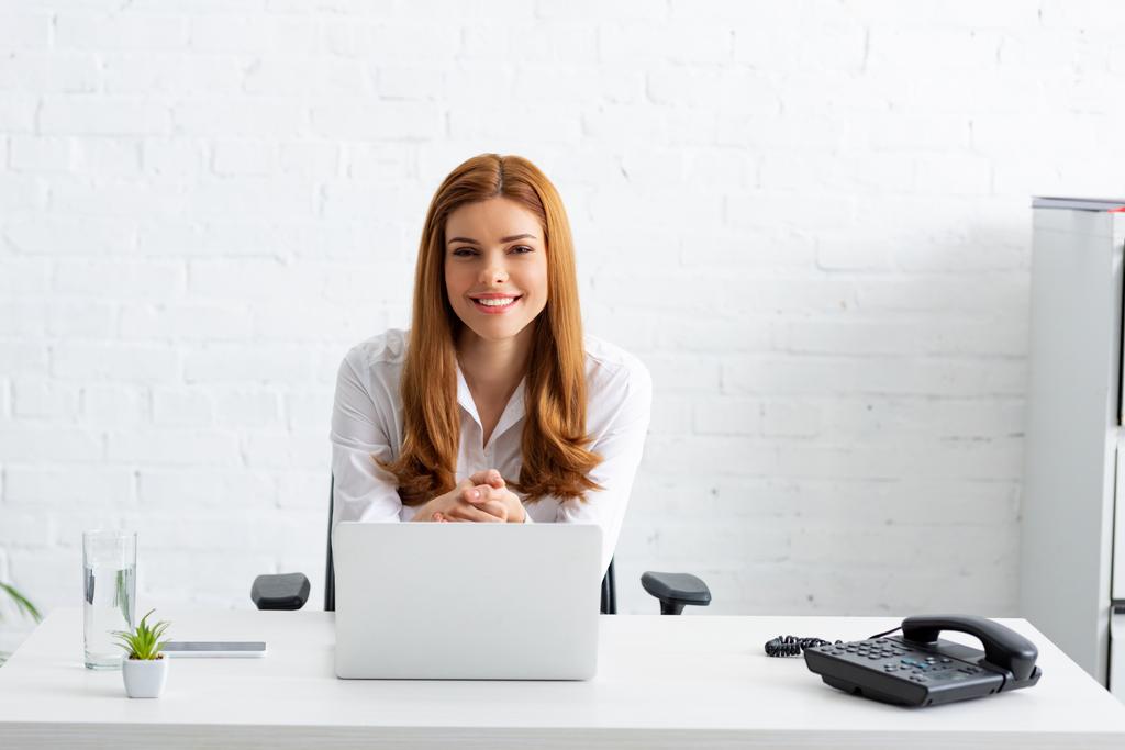 Επιτυχημένη επιχειρηματίας χαμογελώντας στην κάμερα κοντά στο φορητό υπολογιστή και το τηλέφωνο στο τραπέζι - Φωτογραφία, εικόνα