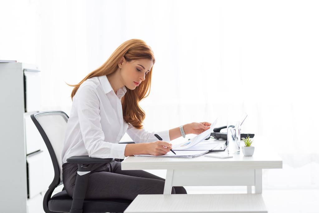 Seitenansicht einer Geschäftsfrau, die mit Papieren und Diagrammen am Bürotisch arbeitet  - Foto, Bild