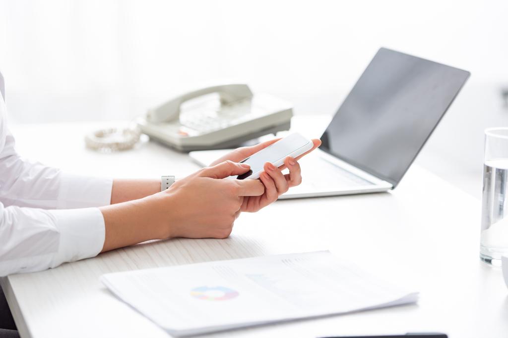 Обрезанный вид деловой женщины с помощью смартфона рядом с документами и ноутбуком на столе
 - Фото, изображение