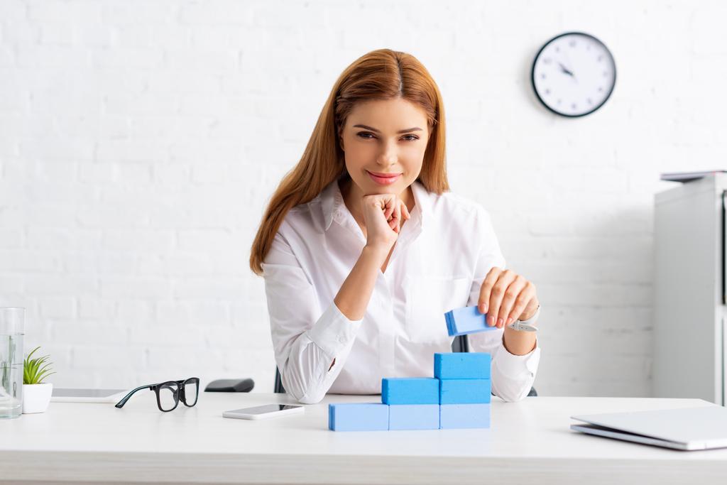 Successful businesswoman smiling at camera while stacking marketing pyramid from building blocks on table - 写真・画像