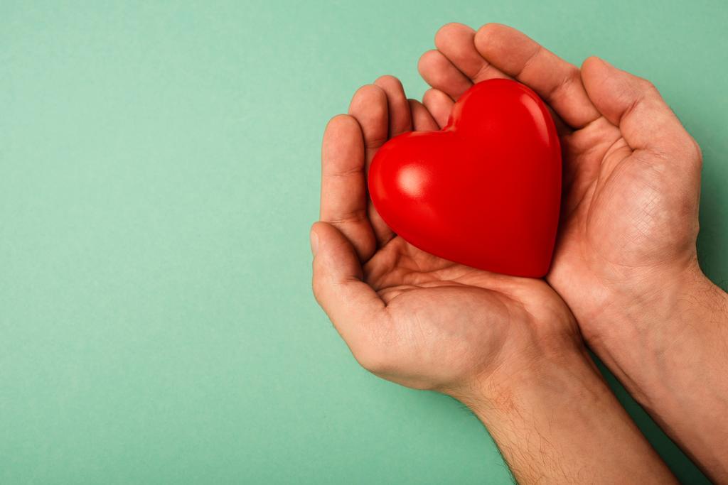 Gekropte weergave van decoratieve rode hart in de handen van de mens op groene achtergrond, wereld gezondheidsdag concept - Foto, afbeelding