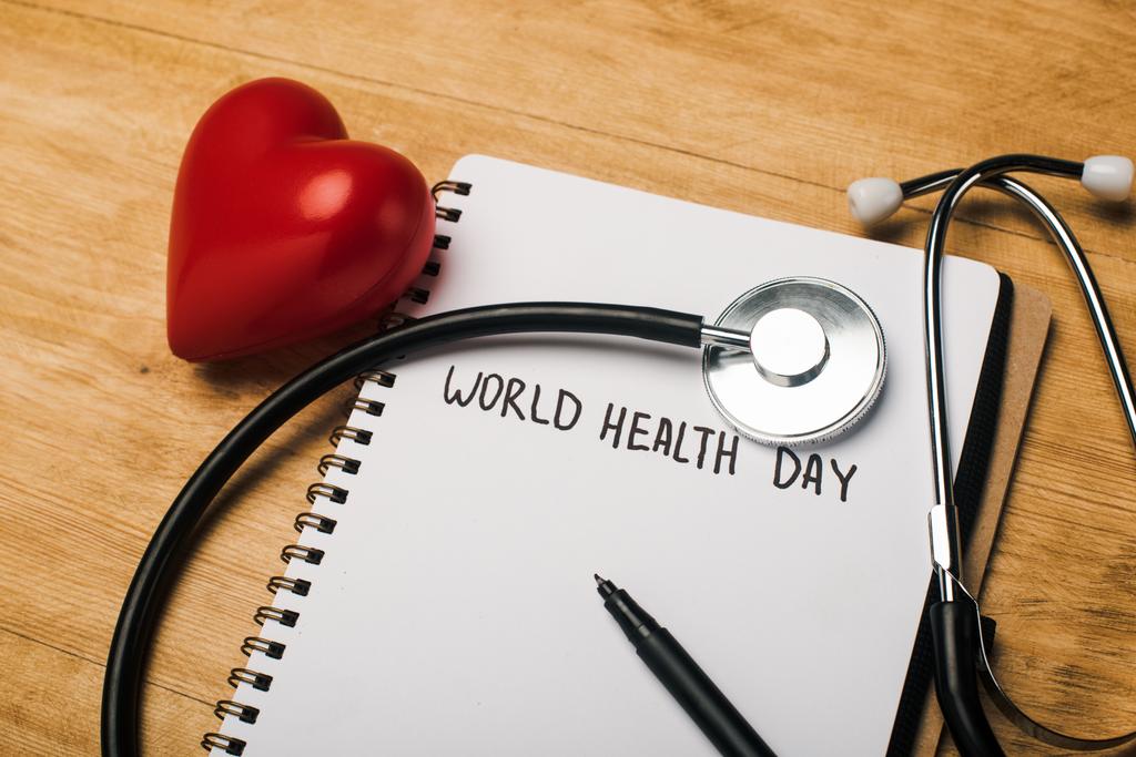 Stetoskop, dekoratif kalp, defter üzerine kalem, tahtadan arka planda Dünya Sağlık Günü yazıları. - Fotoğraf, Görsel