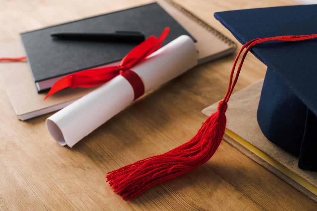 ノートブック、ペン、卒業証書と木製の背景に赤いタッセル付き卒業キャップの選択的な焦点 - 写真・画像