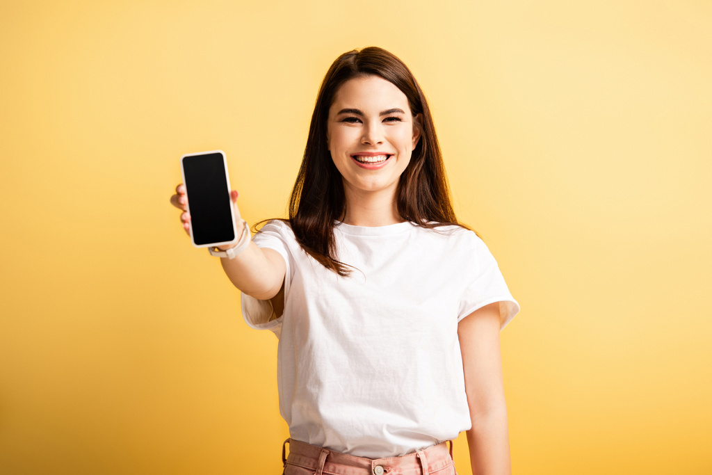 happy girl showing smartphone with blank screen while smiling at camera on yellow background - Photo, Image