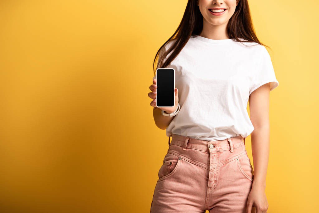 Gülümseyen kızın sarı arkaplanda boş ekranlı akıllı telefon göstermesi. - Fotoğraf, Görsel
