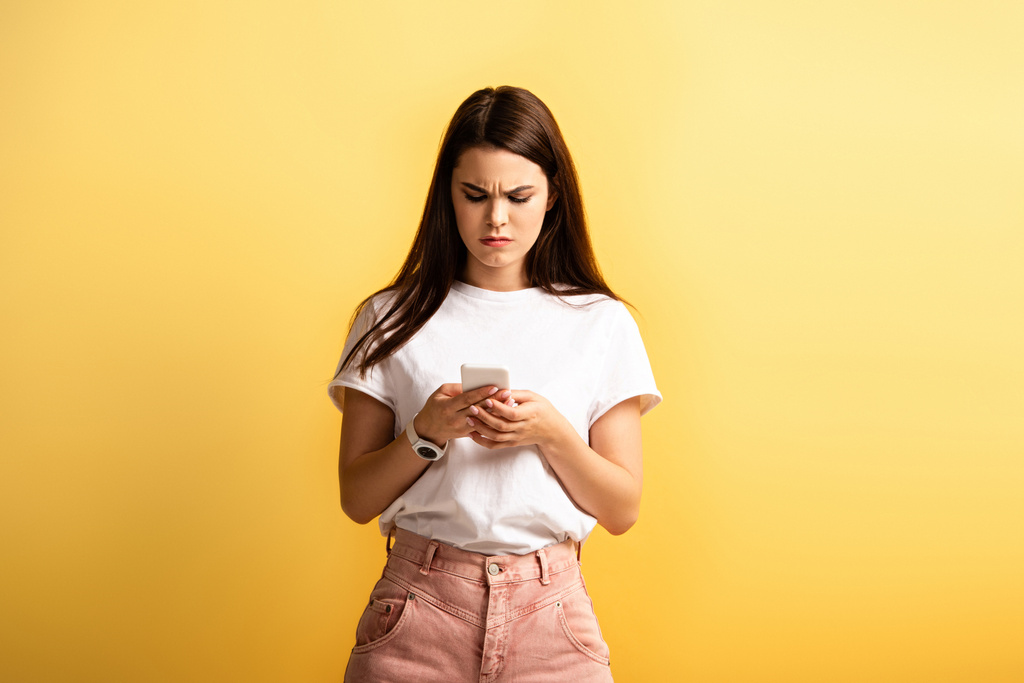 niezadowolona dziewczyna marszczenie brwi podczas wiadomości na smartfonie odizolowany na żółto - Zdjęcie, obraz