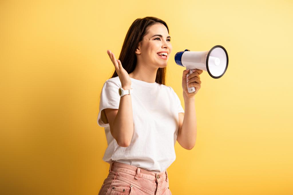 happy girl speaking in megaphone while standing with open arm and looking away on yellow background - Photo, Image