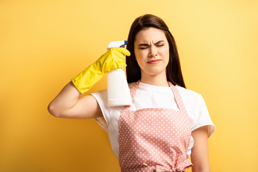 młoda gospodyni domowa w fartuchu i gumowych rękawiczkach imitujących samozabijanie z butelką z rozpylaczem z zamkniętymi oczami na żółtym tle - Zdjęcie, obraz