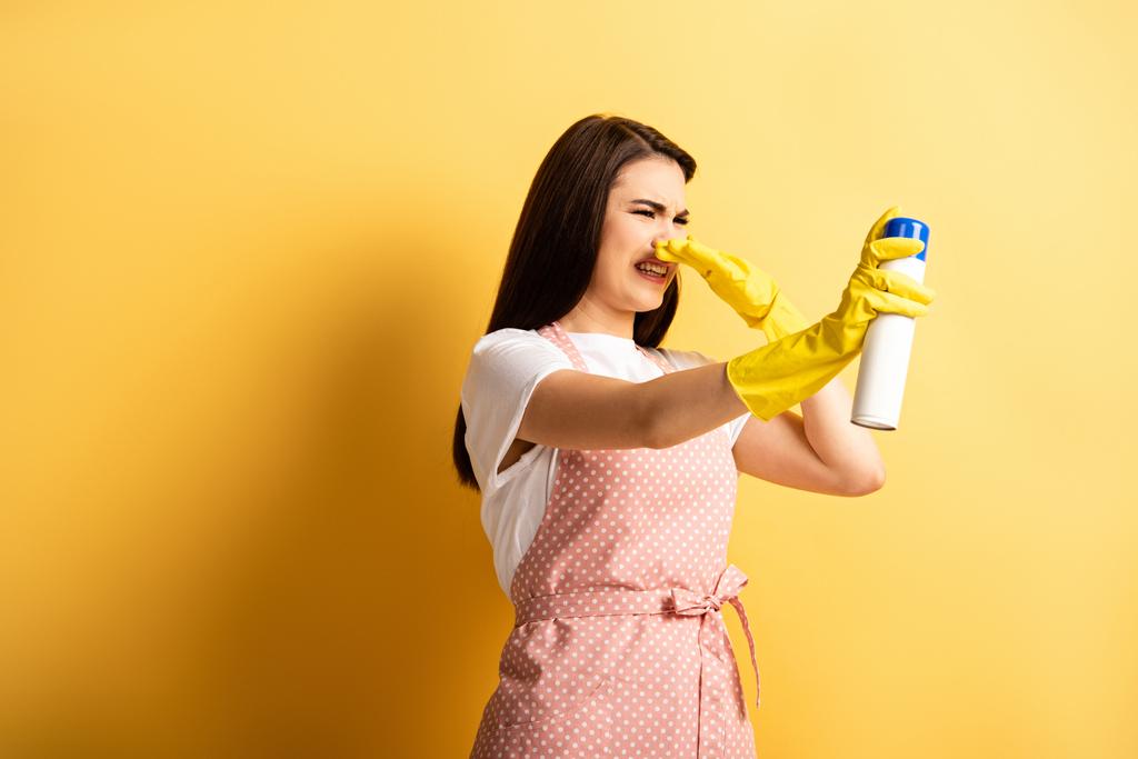 niezadowolona gospodyni domowa w fartuchu i gumowych rękawiczkach zatykająca wąż ręką podczas natryskiwania odświeżacza powietrza na żółtym tle - Zdjęcie, obraz