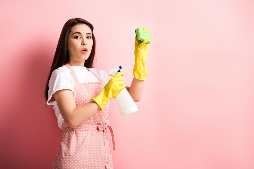 surprise femme au foyer en tablier et gants en caoutchouc tenant bouteille de pulvérisation et éponge sur fond rose
 - Photo, image