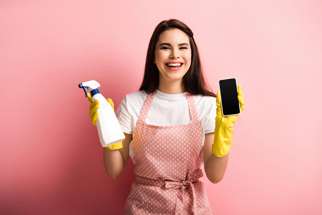 χαρούμενη νοικοκυρά με ποδιά και λαστιχένια γάντια που δείχνει smartphone με λευκή οθόνη ενώ κρατά μπουκάλι σπρέι σε ροζ φόντο - Φωτογραφία, εικόνα