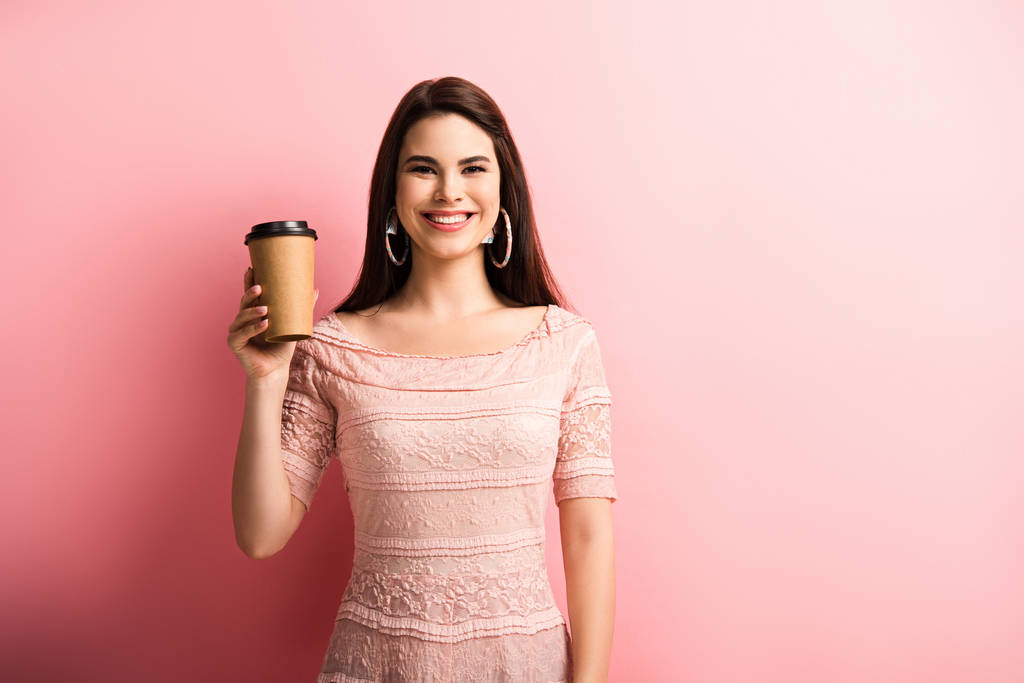 веселая девушка улыбается в камеру, держа кофе, чтобы пойти на розовый фон
 - Фото, изображение