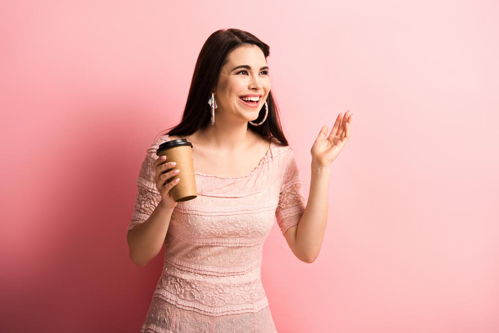 glückliches Mädchen, das wegschaut und lächelt, während es Kaffee auf rosa Hintergrund hält - Foto, Bild