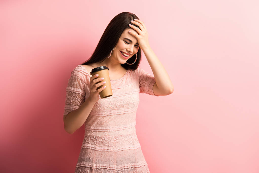 ευτυχισμένο κορίτσι αγγίζοντας το κεφάλι, ενώ κρατώντας καφέ για να πάει σε ροζ φόντο - Φωτογραφία, εικόνα