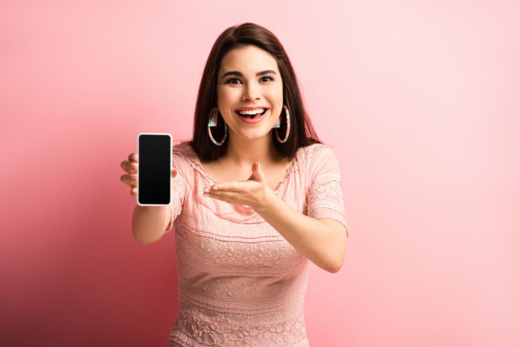 взволнованная девушка указывает на смартфон с пустым экраном на розовом фоне
 - Фото, изображение