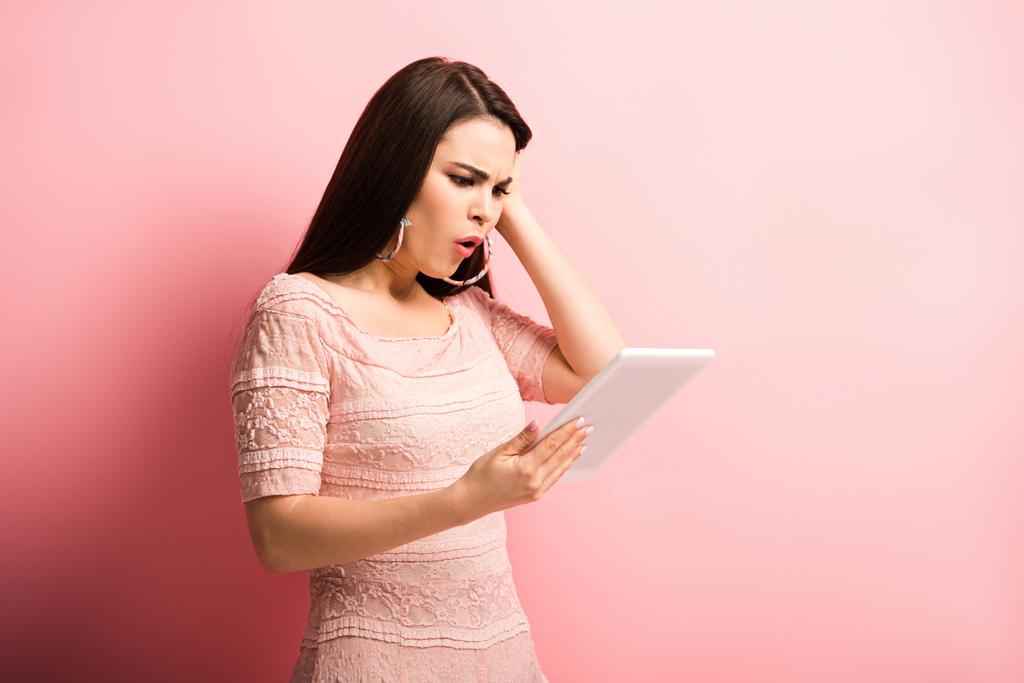 chica conmocionada tocando el cabello mientras mira la tableta digital sobre fondo rosa
 - Foto, imagen