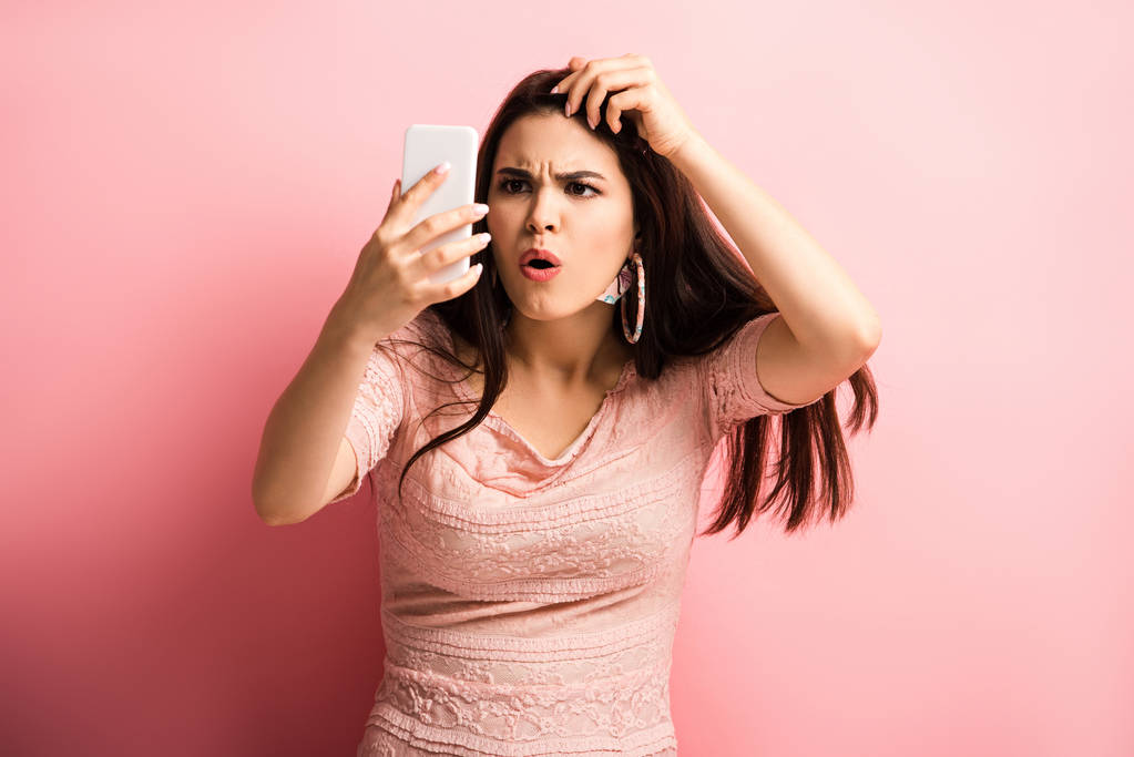 шокированная девушка касаясь волос, глядя на смартфон на розовом фоне
 - Фото, изображение