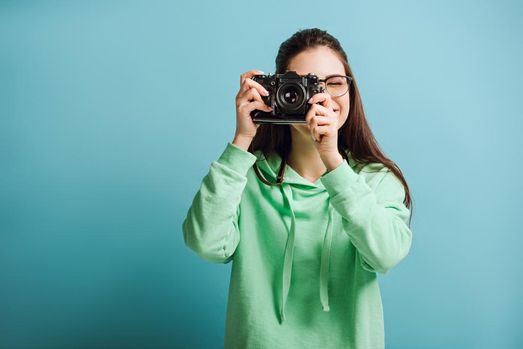 hymyilevä valokuvaaja vihreässä hupparissa ottamassa valokuvaa digitaalisella kameralla sinisellä taustalla
 - Valokuva, kuva