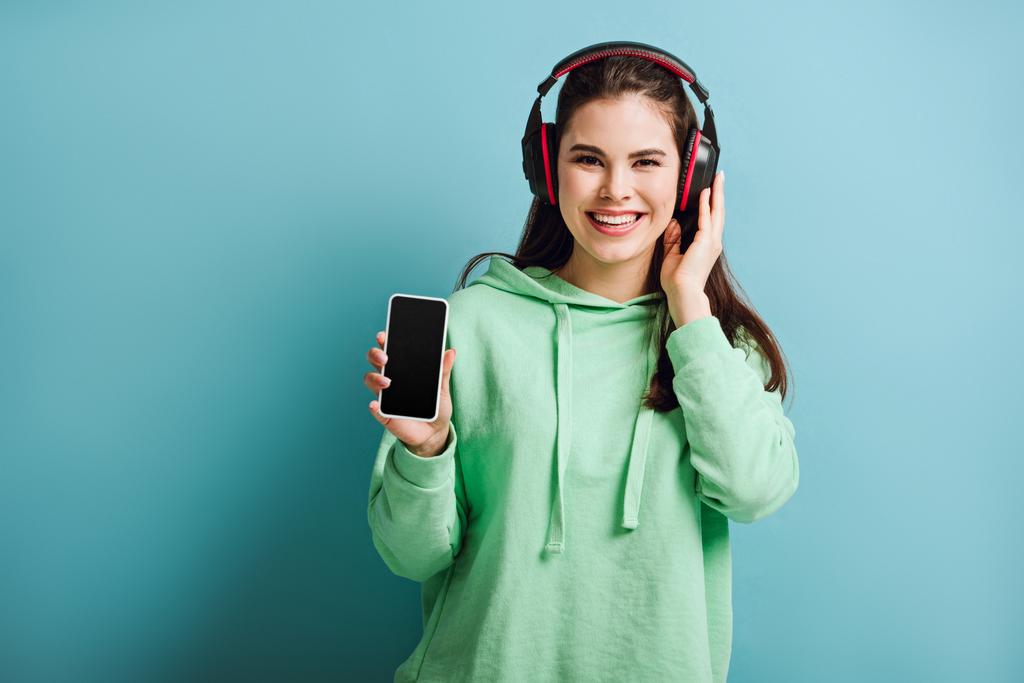 menina alegre em fones de ouvido sem fio olhando para a câmera ao mostrar smartphone com tela em branco no fundo azul
 - Foto, Imagem
