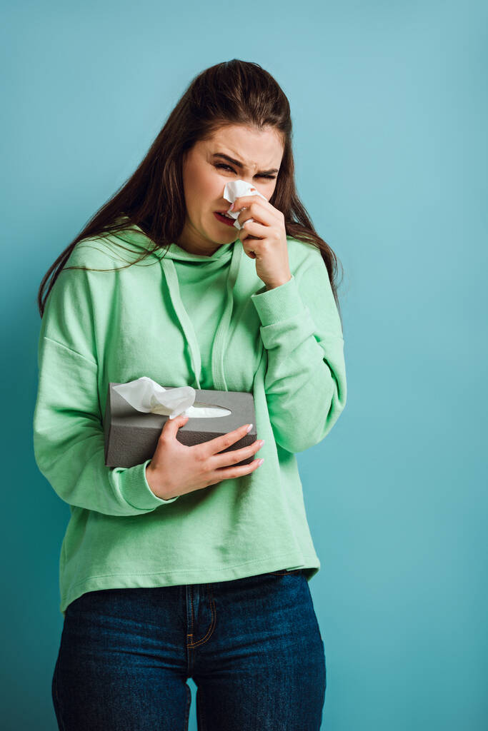 niña enferma limpiando la nariz con servilleta de papel sobre fondo azul
 - Foto, Imagen